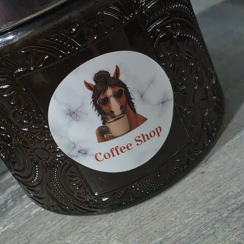 A dark embossed-glass candle jar with a round Coffee Shop label: a horse in a messy bun and heart sunglasses, sipping a coffee.