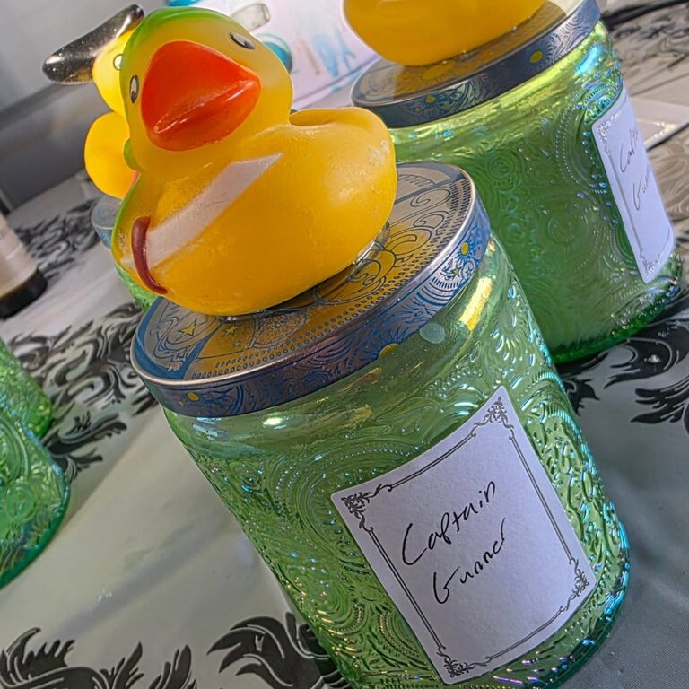 A yellow rubber duck sitting on the lid of a green carved-glass candle jar with a handwritten label reading Captain Gunner.